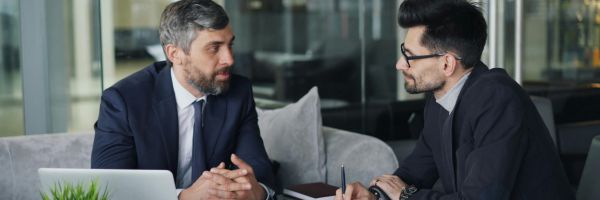 Two men in business attire sit across from each other on a couch, engaged in conversation with a laptop and notebook on the table between them. From Lawrence Law Firm New Jersey Family Lawyer.