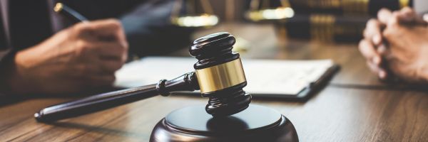 A wooden gavel rests on a desk with two people seated in the background, one writing on a document. From Lawrence Law Firm New Jersey Family Lawyer.