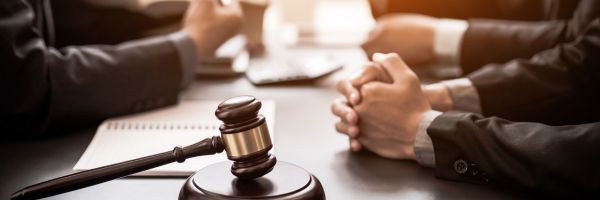 A close-up of a gavel on a table with three people in business suits having a discussion, hands folded, and a notebook in view. From Lawrence Law Firm New Jersey Family Lawyer.