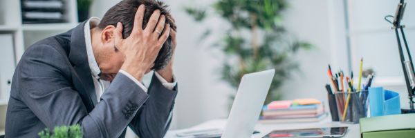 A man in a suit sits at a desk with his head in his hands, appearing stressed or frustrated, with a laptop and office supplies in front of him. From Lawrence Law Firm New Jersey Family Lawyer.