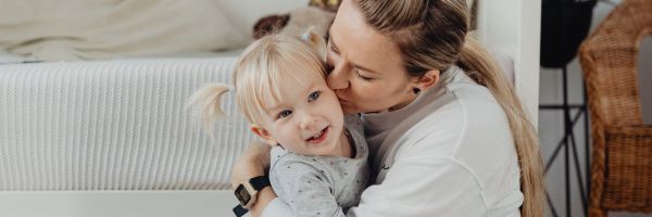 A single parent hugs and kisses a young child with blonde hair in a room with a bed and wicker basket in the background, capturing a tender moment during the holiday season. From Lawrence Law Firm New Jersey Family Lawyer.