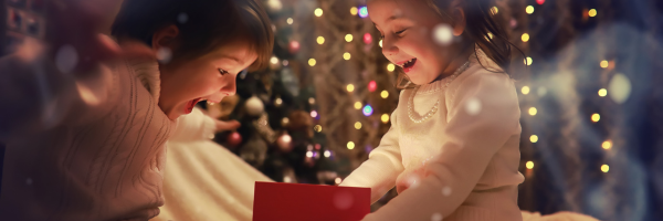 Two young children sit in front of a decorated Christmas tree, smiling and looking excited as they open a red gift box. This joyful moment captures the magic of the holiday season for a single parent family, surrounded by festive lights. From Lawrence Law Firm New Jersey Family Lawyer.