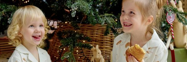 Two young children in pajamas sit in front of a decorated Christmas tree during the holiday season, one holding a pastry, capturing a warm moment for their single parent. From Lawrence Law Firm New Jersey Family Lawyer.
