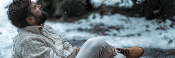 A single parent in a thick beige sweater and tan boots sits on snowy ground outdoors, leaning against a tree with a thoughtful expression during the holiday season. From Lawrence Law Firm New Jersey Family Lawyer.