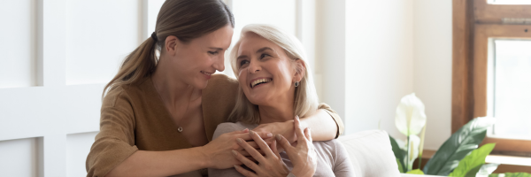 Two women sit on a couch, smiling at each other. One woman, a single parent, wraps her arms around the other in a warm embrace. A window and potted plant highlight the cozy holiday season backdrop. From Lawrence Law Firm New Jersey Family Lawyer.