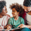 A child sits between two adults, smiling while one adult holds an open book. All three are engaged and looking at each other, enjoying a warm family moment—just the kind of connection Jeralyn Lawrence values. From Lawrence Law Firm New Jersey Family Lawyer.