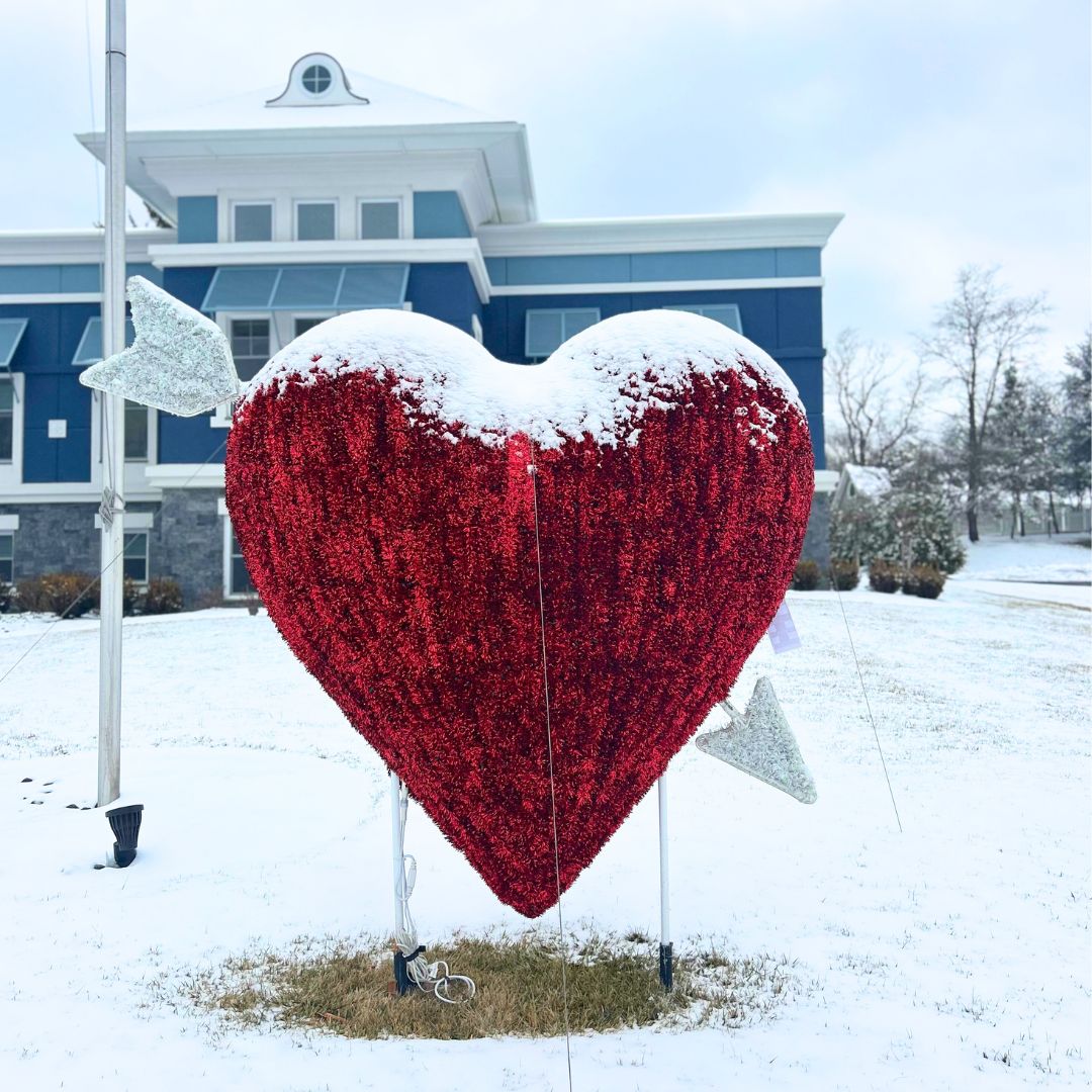 A large red glittery heart decoration with silver arrows and a layer of snow stands outside on a snowy lawn, welcoming visitors to Our Firm in front of a blue building. From Lawrence Law Firm New Jersey Family Lawyer.