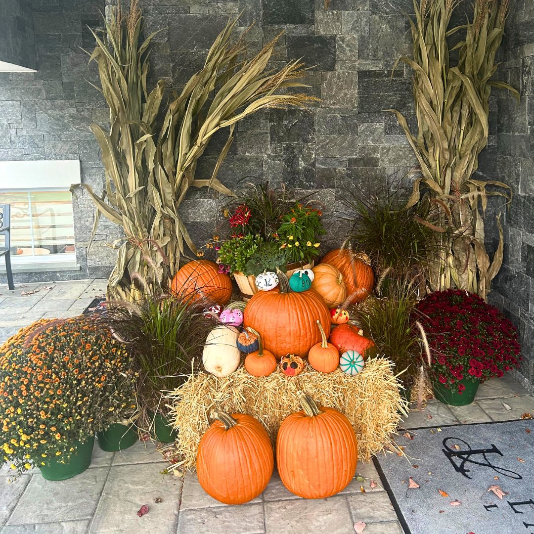 An arrangement of pumpkins, painted gourds, hay bales, dried cornstalks, and potted mums by Our Firm decorates a stone patio. From Lawrence Law Firm New Jersey Family Lawyer.