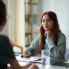 A young woman sits at a desk, attentively listening to another person in an office setting with papers and a glass of water in front of her—perhaps discussing a case with Jeralyn Lawrence. From Lawrence Law Firm New Jersey Family Lawyer.