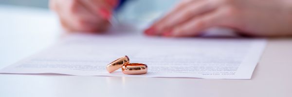 Two gold wedding rings rest on a legal document, symbolizing the importance of prenuptial agreements as a person signs paperwork in the background. From Lawrence Law Firm New Jersey Family Lawyer.