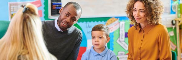 A young boy sits between two adults, likely his parents, speaking with a teacher in a colorful classroom setting—reflecting discussions often seen in New Jersey custody cases focused on the best interests of the child. From Lawrence Law Firm New Jersey Family Lawyer.