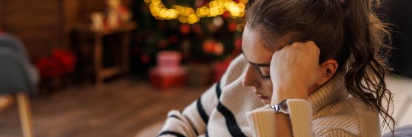 A woman in a striped sweater sits indoors with her head resting on her hand, looking down—capturing the reflective mood often seen during Divorce Month, as blurred Christmas decorations and a lit tree glow in the background. From Lawrence Law Firm New Jersey Family Lawyer.