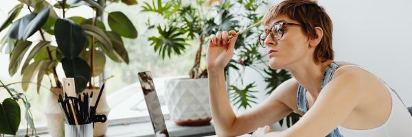 A person wearing glasses sits at a desk with a laptop, holding a pencil and looking thoughtful, surrounded by potted plants—perhaps reflecting on Divorce Month and the January divorce trends that spark new beginnings. From Lawrence Law Firm New Jersey Family Lawyer.
