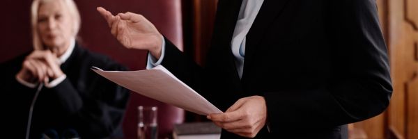 A person in a suit holds papers and speaks in a courtroom during Divorce Month, while another person sits in the background wearing judicial robes. From Lawrence Law Firm New Jersey Family Lawyer.