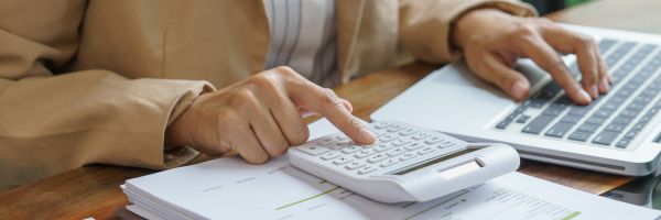 Person in a beige jacket uses a calculator on top of documents, typing on a laptop—perhaps preparing to file for divorce in NJ and organize finances for the New Year. From Lawrence Law Firm New Jersey Family Lawyer.