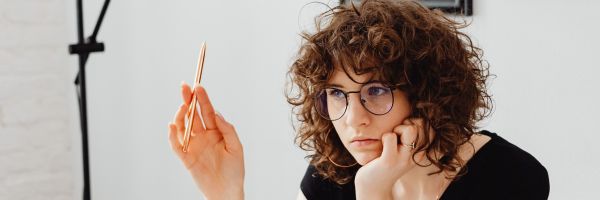Person with curly hair and glasses sits at a desk, holding a pen and looking thoughtful about how to file for divorce in NJ, their chin resting on their hand. From Lawrence Law Firm New Jersey Family Lawyer.