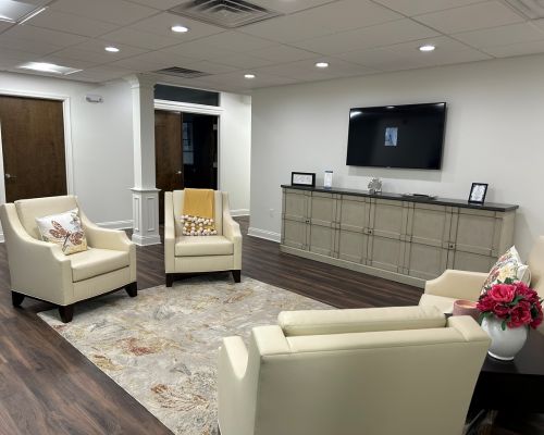 A welcoming waiting room in Our Firm features four cream armchairs around a rug, a cabinet with framed photos, a vase of flowers, and a wall-mounted TV in a clean, well-lit space. From Lawrence Law Firm New Jersey Family Lawyer.