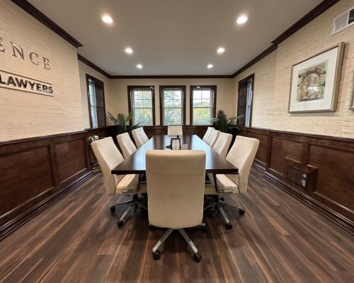 A conference room in Our Firm features a long table, ten white chairs, wood-paneled walls, framed art, and large windows letting in abundant natural light. From Lawrence Law Firm New Jersey Family Lawyer.