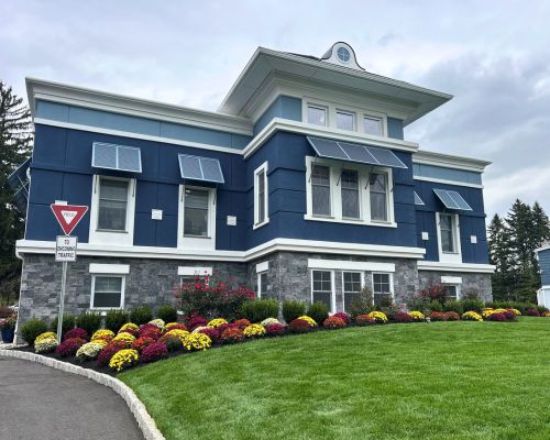 Our Firm occupies a two-story blue building with large windows, gray stone accents, solar panels, landscaped bushes, and colorful flowers, with a yield sign on the left side. From Lawrence Law Firm New Jersey Family Lawyer.
