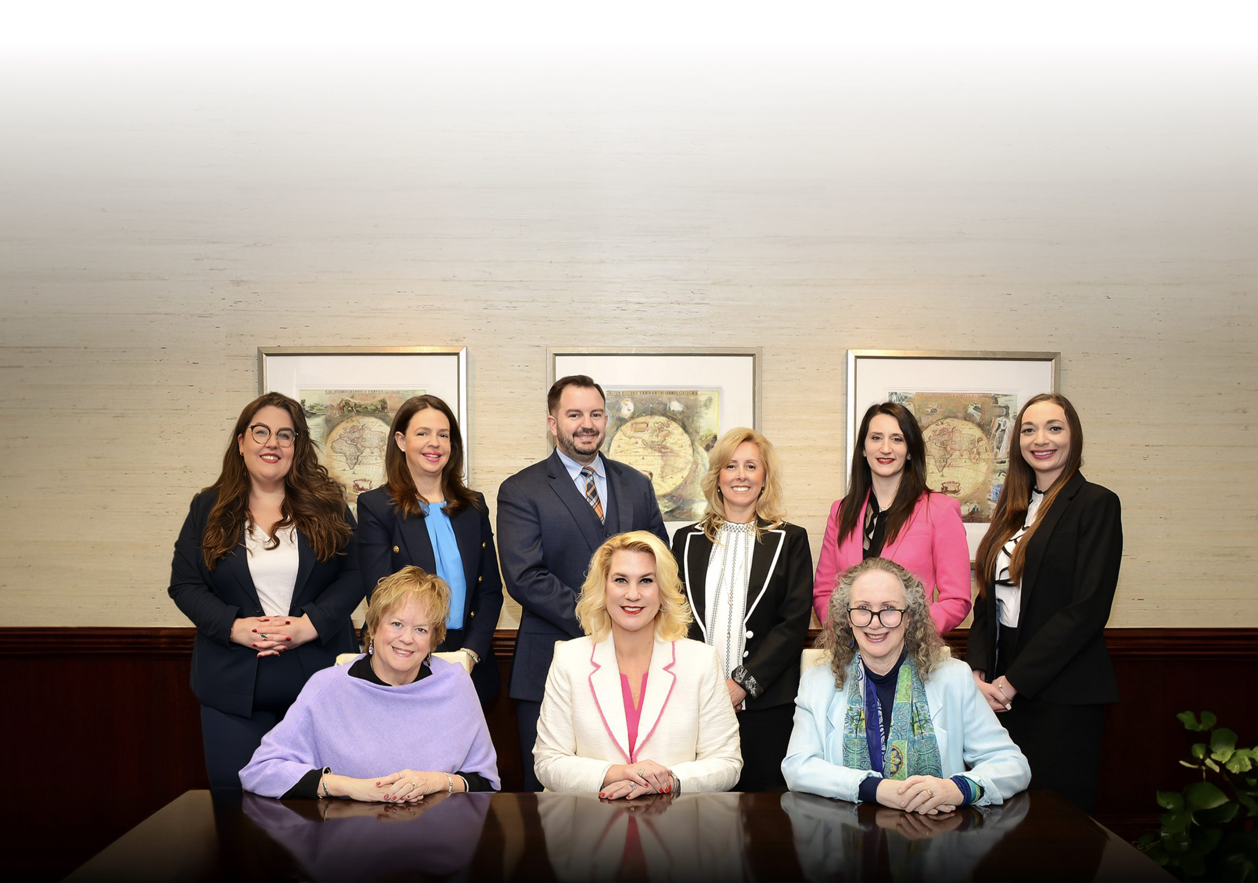 Nine professionally dressed people, six standing and three seated, pose for a group photo in an office setting with two framed maps on the wall behind them. From Lawrence Law Firm New Jersey Family Lawyer.