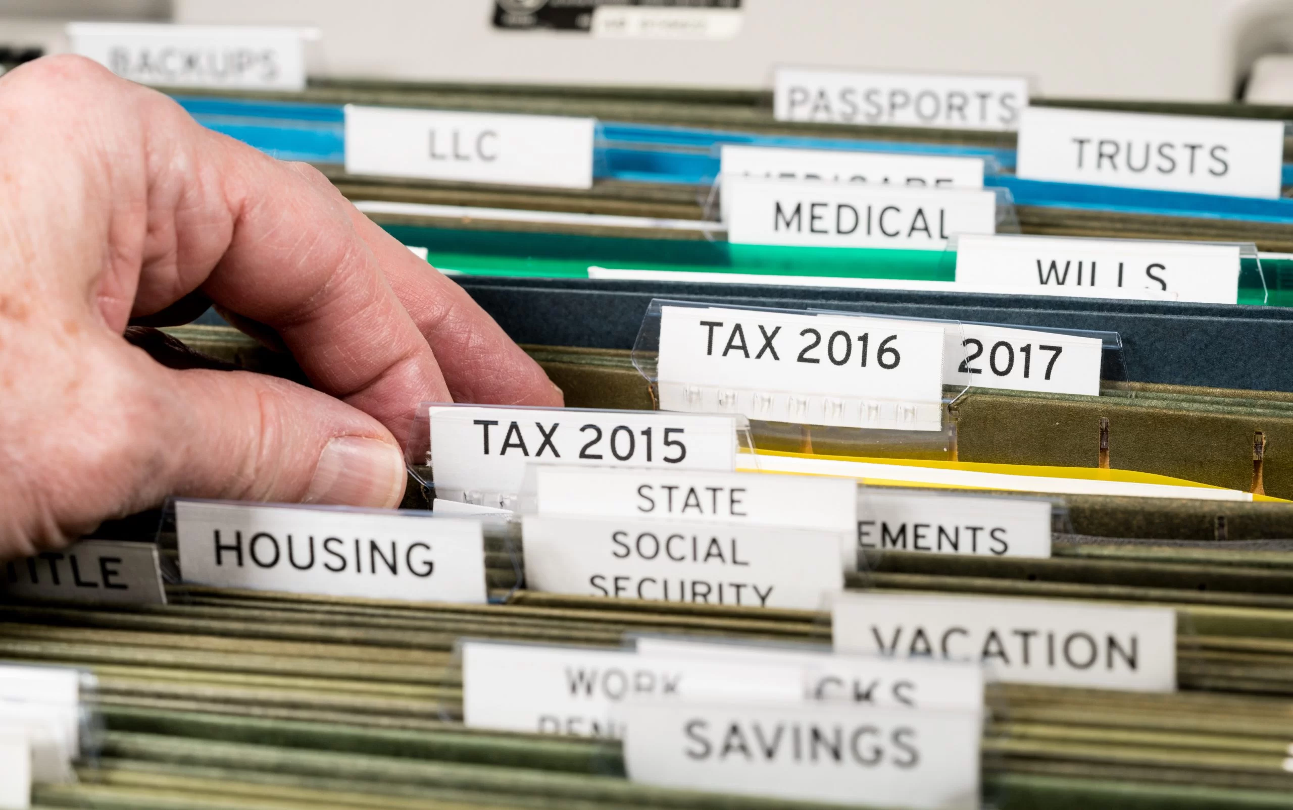 A hand selects a file labeled “TAX 2015” from a drawer containing folders with labels such as “HOUSING,” “SAVINGS,” “WILLS,” and “MEDICAL”—perfect for organizing a Year-End Divorce Checklist. From Lawrence Law Firm New Jersey Family Lawyer.
