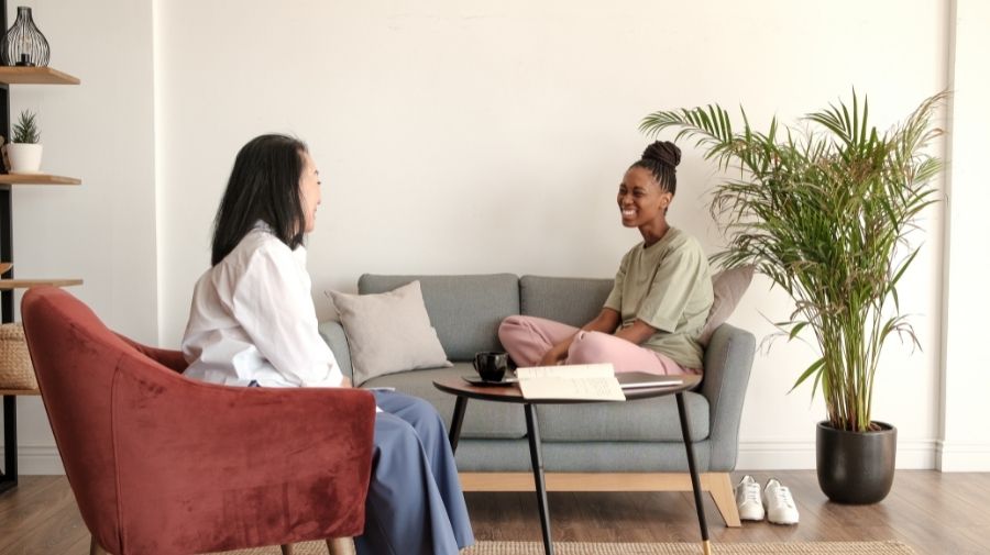 Two women sit facing each other in a living room, one on a sofa and one on a chair, discussing their year-end divorce checklist with a table and potted plant nearby. From Lawrence Law Firm New Jersey Family Lawyer.