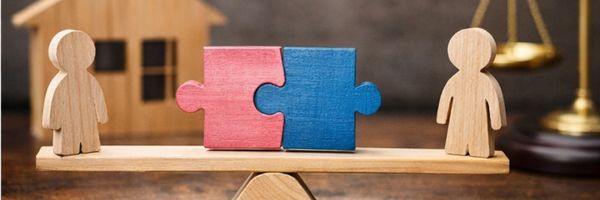 Two wooden human figures stand on each end of a balanced scale, with red and blue puzzle pieces joined in the center; a wooden house and scales of justice are in the background. From Lawrence Law Firm New Jersey Family Lawyer.