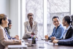 A group of six people sit around a conference table having a meeting, with one person standing and speaking to the group in a modern office setting. From Lawrence Law Firm New Jersey Family Lawyer.