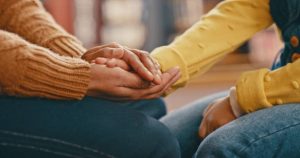 Two people sit side by side holding hands, one wearing a brown sweater and the other a yellow one, conveying comfort or support. From Lawrence Law Firm New Jersey Family Lawyer.