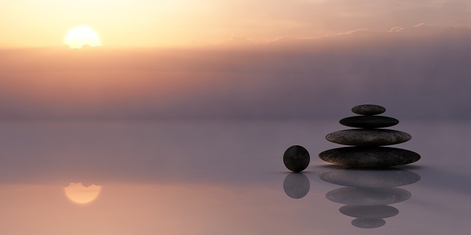 Smooth stones stacked on a glassy surface reflect in water at sunset, with a soft orange sky and sun partially visible above the horizon. From Lawrence Law Firm New Jersey Family Lawyer.