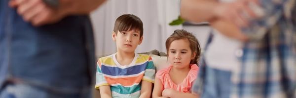 Two children sit on a couch with serious expressions while two adults stand in the foreground, partially out of focus. From Lawrence Law Firm New Jersey Family Lawyer.
