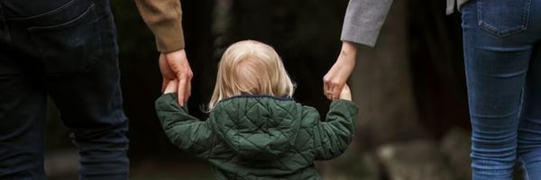 A small child in a green jacket holds hands with two adults, one on each side, as they walk together outdoors. From Lawrence Law Firm New Jersey Family Lawyer.