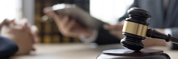 A wooden judge’s gavel rests on a desk, with two people in business attire discussing documents in the background. From Lawrence Law Firm New Jersey Family Lawyer.