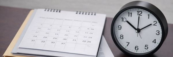 A black analog clock showing 11:55 is placed next to an open desk calendar with visible dates and papers underneath. From Lawrence Law Firm New Jersey Family Lawyer.