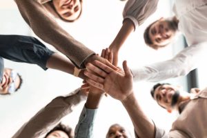 A group of people standing in a circle stack their hands together in the center, viewed from below, symbolizing teamwork and unity. From Lawrence Law Firm New Jersey Family Lawyer.