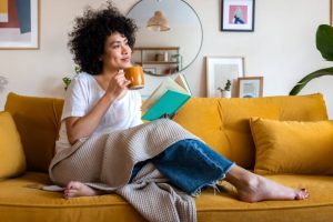 A person sits on a yellow sofa with a blanket, holding a mug in one hand and an open book in the other. The room is decorated with framed art and plants. From Lawrence Law Firm New Jersey Family Lawyer.